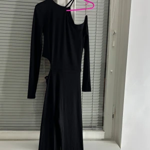 Svart oneshoulder jumpsuit - Snygg svart oneshoulder jumpsuit med lång ärm och ribbad textur. Den har en unik design med en öppen axel och knytning i midjan. Perfekt för en stilren look. Aldrig använt