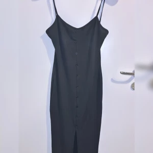 Svart långklänning med knappar - Elegant svart långklänning med smala axelband och knappar längs framsidan. Klänningen har en stilren slits nedtill som ger en snygg detalj. Perfekt för en chic look.