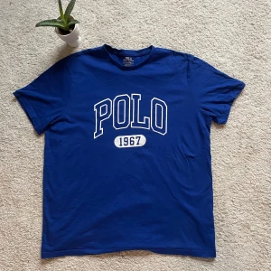 Blå t-shirt från Ralph Lauren - Säljer en blå t-shirt från Ralph Lauren med texten 'POLO 1967' i vitt på framsidan. T-shirten har en klassisk rund hals och korta ärmar. Perfekt för en avslappnad stil. 🌟
