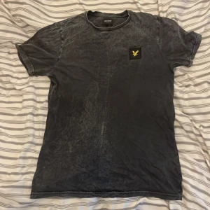 Grå t-shirt från Lyle & Scott - Säljer en grå t-shirt från Lyle & Scott med korta ärmar och ett gult broderat emblem på bröstet. Perfekt för en avslappnad stil. T-shirten har en tvättad look som ger den en unik känsla.