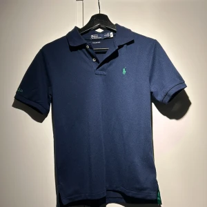 Blå pikétröja från Ralph Lauren - Säljer en stilren blå pikétröja från Ralph Lauren med grönt broderat logotyp på bröstet och ärmen. Tröjan har korta ärmar och en klassisk krage med knappar. Perfekt för en avslappnad men ändå snygg look.