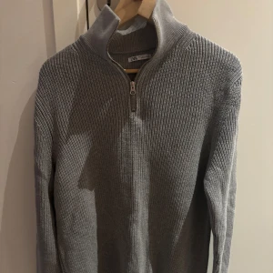 Grå stickad tröja från Zara - Säljer en stilren grå stickad tröja från Zara med dragkedja och hög krage. Perfekt för kyligare dagar med sin mjuka och bekväma passform. Tröjan har långa ärmar och en ribbad struktur som ger en klassisk look.