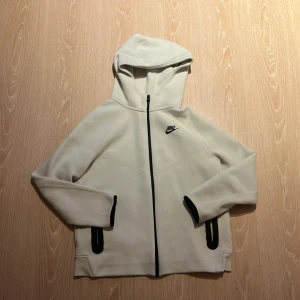 Vit hoodie från Nike - Säljer en stilren vit hoodie från Nike med svart dragkedja och logga. Den har en praktisk huva och långa ärmar. Perfekt för en sportig look eller avslappnade dagar.