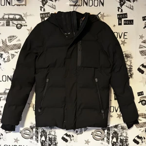 Svart dunjacka från Jack and Jones  - Säljer en stilren svart dunjacka från Jack and Jones. Jackan har en praktisk huva och dragkedja framtill. Insidan är fodrad med ett snyggt mönster och har orange detaljer. Perfekt för kyliga dagar. Aldrig använd.