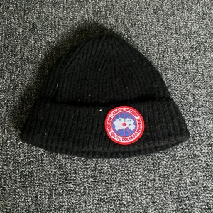 Svart stickad mössa från Canada Goose - Snygg svart stickad mössa från Canada Goose med ett broderat märke framtill. Perfekt för kalla dagar och ger en stilren look. Mössan är ribbad och har en klassisk design.