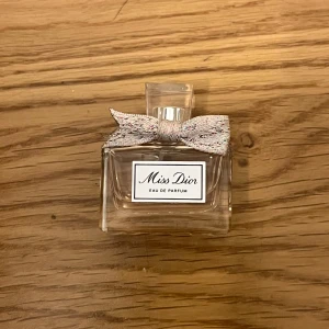 Miss Dior Eau de Parfum mini - Säljer en elegant Miss Dior Eau de Parfum i en stilren glasflaska med en söt rosett på korken. Flaskan rymmer 5 ml och har en lyxig känsla. Perfekt för den som älskar en klassisk och tidlös doft. DEN ÄR LITE ANVÄND, SOM DET SYNS PÅ BILD!!