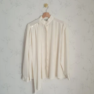 Beige skir vintage blus - 🧁FÖRST TILL KVARN!🧁 En elegant krämvit vintage blus. Skirt i materialet med en en hängande sjal vid nyckelbenet. Kan hängas runt axeln som en lös krage eller fram fritt. Den är avtagbar. På tredje bild syns hur detaljerad tyget är som invävd sidentråd som glänser i olika vinklar. En äkta ögonskatt😍 Strl s/m.