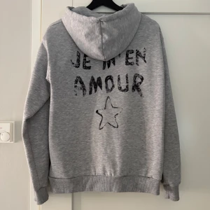 Grå hoodie med text och stjärna - Säljer en grå hoodie med texten 'Je m'en amour' och en stjärna tryckt på både fram- och baksidan. Den har en justerbar huva med snören och ribbade muddar vid ärmslut och nederkant. Perfekt för en avslappnad stil.