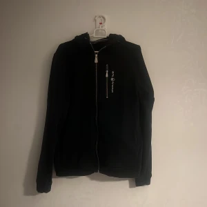 Svart jacka från Sail Racing - Snygg svart jacka från Sail Racing med dragkedja framtill och en extra dragkedja på bröstet. Jackan har en huva och är långärmad, perfekt för en stilren look. Logotypen är tryckt på bröstet och i nacken. Den är också en ganska gammal model så många de är inte många som har den. Så den är perfekt för folk som vill sticka ut