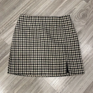 Rutig kjol från Kappahl - Snygg rutig kjol från Kappahl i svart och beige. Kjolen har en liten slits framtill och är perfekt för en stilren look. Den är kort och har en klassisk passform.