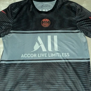 Svart PSG fotbollströja från Nike - Snygg svart PSG fotbollströja från Nike med röd logga och Dri-FIT material. Tröjan har korta ärmar och ett randigt mönster.