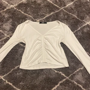 Vit långärmad topp från Bik Bok - Säljer en stilren vit långärmad topp från Bik Bok. Toppen har en v-ringad design med rynkdetaljer framtill som ger en snygg look. Perfekt för att matcha med jeans eller kjol. Dock lite nopprig men syns inte.