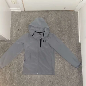 Grå vindjacka från Under Armour - Säljer en grå vindjacka från Under Armour med huva och dragkedja. Jackan har en stilren design med en diskret logotyp på bröstet. Perfekt för blåsiga dagar.