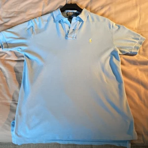 Ljusblå pikétröja från Polo Ralph Lauren - Säljer en stilren ljusblå pikétröja från Polo Ralph Lauren. Tröjan är kortärmad och har en klassisk krage med knappar. Den är tillverkad i 100% bomull och har det ikoniska broderade logotypen på bröstet. Perfekt för en avslappnad och snygg look.