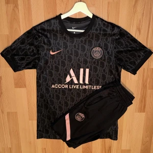 Psg Svart fotbollströja och shorts från Nike - Snygg svart fotbollströja och shorts från Nike med rosa detaljer och PSG-logga. Tröjan har korta ärmar och ett diskret mönster. Perfekt för fotbollsfans som vill ha stil på planen.