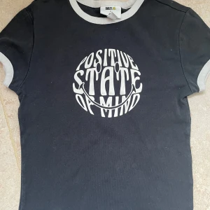 Svart t-shirt med tryck från H&M stl S  - Snygg svart t-shirt från H&M med ett coolt tryck 'Positive State of Mind' i vitt. T-shirten har kontrasterande vita kanter vid hals och ärmar, vilket ger en retro känsla. Perfekt för en avslappnad stil.