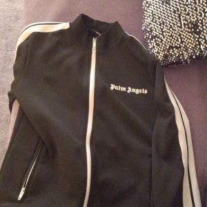 Svart track jacket från Palm Angels - Säljer en svart track jacket från Palm Angels med vit dragkedja och vita ränder längs ärmarna. Jackan har en broderad logotyp på bröstet och en hög krage. Perfekt för killar