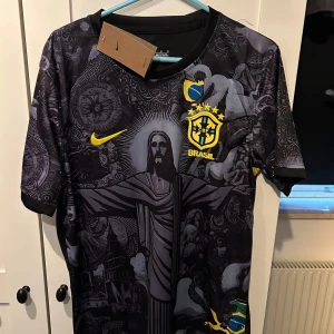 Svart Brasilien fotbollströja från Nike - Snygg svart fotbollströja från Nike med Brasilien-tema. Tröjan har ett detaljerat mönster med Kristusstatyn och andra symboler. Den har korta ärmar och det ikoniska Nike-märket i gult. Perfekt för fotbollsfans som vill sticka ut!