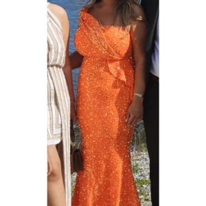 Orange glittrig långklänning - Säljer en fantastisk orange långklänning med glittrande paljetter. Klänningen har en elegant offshoulder-design och en vacker volangdetalj framtill. Perfekt för festliga tillfällen där du vill sticka ut och glänsa. Passar perfekt för sommarens alla fester! 🌟