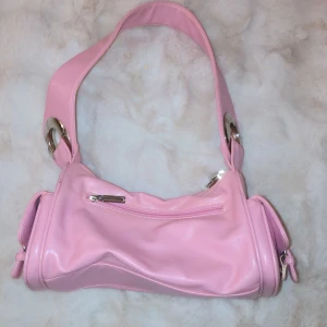 Rosa axelväska med silverdetaljer - Säljer en superfin rosa axelväska i syntetmaterial. Väskan har en cool design med stora silverringar och en praktisk dragkedja framtill. Perfekt för att liva upp vilken outfit som helst! 💖
