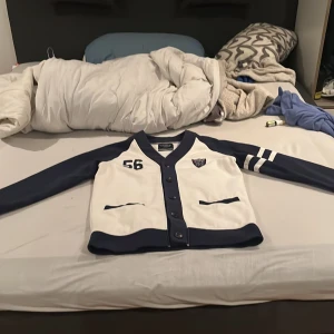 Vit och marinblå kofta från Bondelid - Säljer en snygg vit och marinblå kofta från Bondelid i storlek L. Den har knappar framtill och coola detaljer som ett tryck med siffran 56 och ett Bondelid-märke på bröstet. Perfekt för en avslappnad stil eller en dag på stan. 🧥