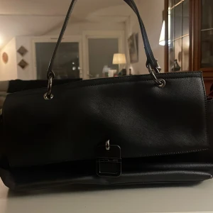 Svart axelväska med röd insida - Snygg svart axelväska i skinn med en elegant design. Väskan har en stilren spänndetalj på framsidan och en lyxig röd insida med dragkedjeficka. Perfekt för både vardag och fest!