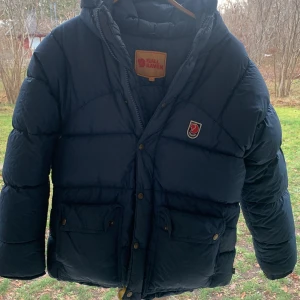 Mörkblå dunjacka från Fjällräven - Säljer en mörkblå dunjacka från Fjällräven  i bra skick. Jackan har en varm och vadderad design med långa ärmar och praktiska fickor framtill. Perfekt för kalla vinterdagar. Den har en dragkedja och knappar för extra skydd mot kyla. Fjällrävens klassiska logga pryder bröstet.