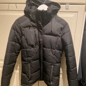 Svart pufferjacka från Clique - Säljer en svart pufferjacka från Clique i storlek S. Jackan har en dragkedja framtill och en praktisk huva. Den är helt ny och aldrig använd. 