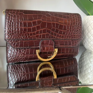 Brun äktaskinn axelväska - Snygg brun axelväska i äktaskinn med gulddetaljer. Väskan har en elegant design med ett stort guldfärgat spänne framtill. Perfekt för både vardag och fest!