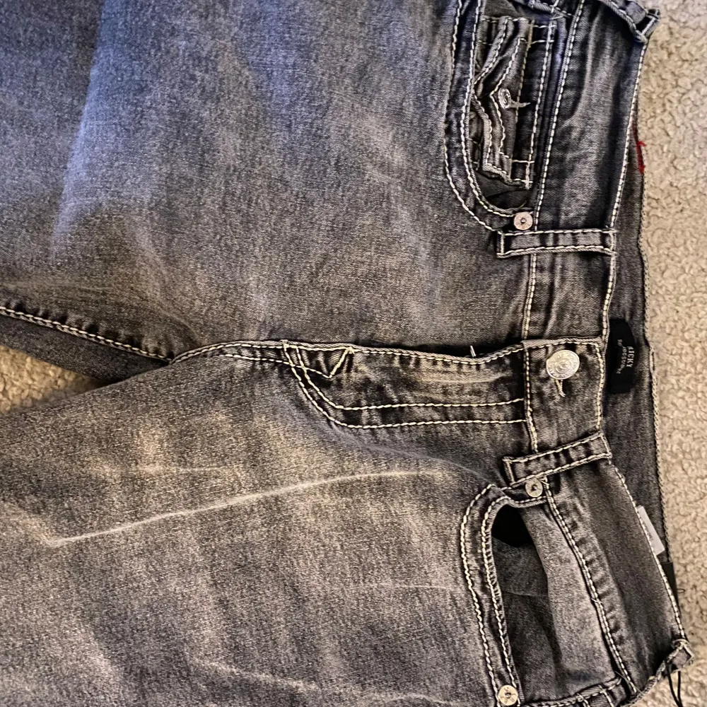 HELT NYA Säljer ett par grå jeans från True Religion i modellen Ricky Big T Flap. De har en straight fit och är i en snygg grå tvätt med slitna detaljer. Bakfickorna har coola sömmar och knappar. Perfekta för en avslappnad stil!. Jeans & Byxor.