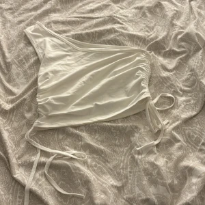 Vit oneshoulder topp - Säljer en stilren vit oneshoulder topp med knytning i sidan. Perfekt för sommarens fester eller en dag på stan. Toppen är i ett mjukt och bekvämt material som sitter snyggt på kroppen.