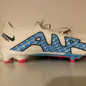 Vita Nike fotbollsskor - Säljer ett par vita Nike fotbollsskor i syntetmaterial med coola blå detaljer och rosa sula. Skorna har snörning och är i mycket bra skick. Perfekta för fotbollsträningen och matcher. Skostorlek EU 43. Dom är bara andvända 5 gånger då jag spelat en handbollscup som är utomhus. skorna kostar 1.400kr