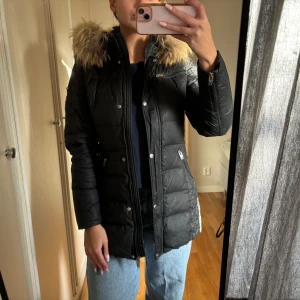 Svart dunjacka från Rockandblue - Säljer en snygg svart dunjacka från Rockandblue med äkta pälskrage. Jackan har en quiltad design och dragkedjor både fram och på fickorna. Perfekt för kalla vinterdagar! Jackan har lite skador på baksidans nedre kant och lite fläckar på insidan av kragen som säkert går bort i tvätt.