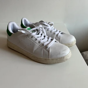 Adidas Stan Smith sneakers - Säljer ett par klassiska Adidas Stan Smith sneakers i vitt med gröna detaljer. Skorna har snörning och är i bra skick med lite slitage på sulan. 