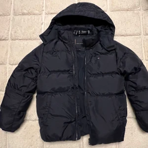 Mörkblå dunjacka från Tommy Hilfiger - Säljer en mörkblå dunjacka från Tommy Hilfiger i mycket bra skick. Köpt 2023 och knappt använd! Jackan har en dragkedja framtill och avtagbar luva. Den är långärmad och har en klassisk pufferstil. Passar kille 160-170 cm.