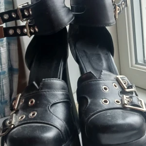 Svarta platåskor med spännen - Säljer ett par coola svarta platåskor i syntetmaterial. De har flera spännen och dragkedja bak för en edgy look. Perfekta för en utekväll eller fest! Skorna har en rejäl klack som är super hög! For my alts and emos out there!