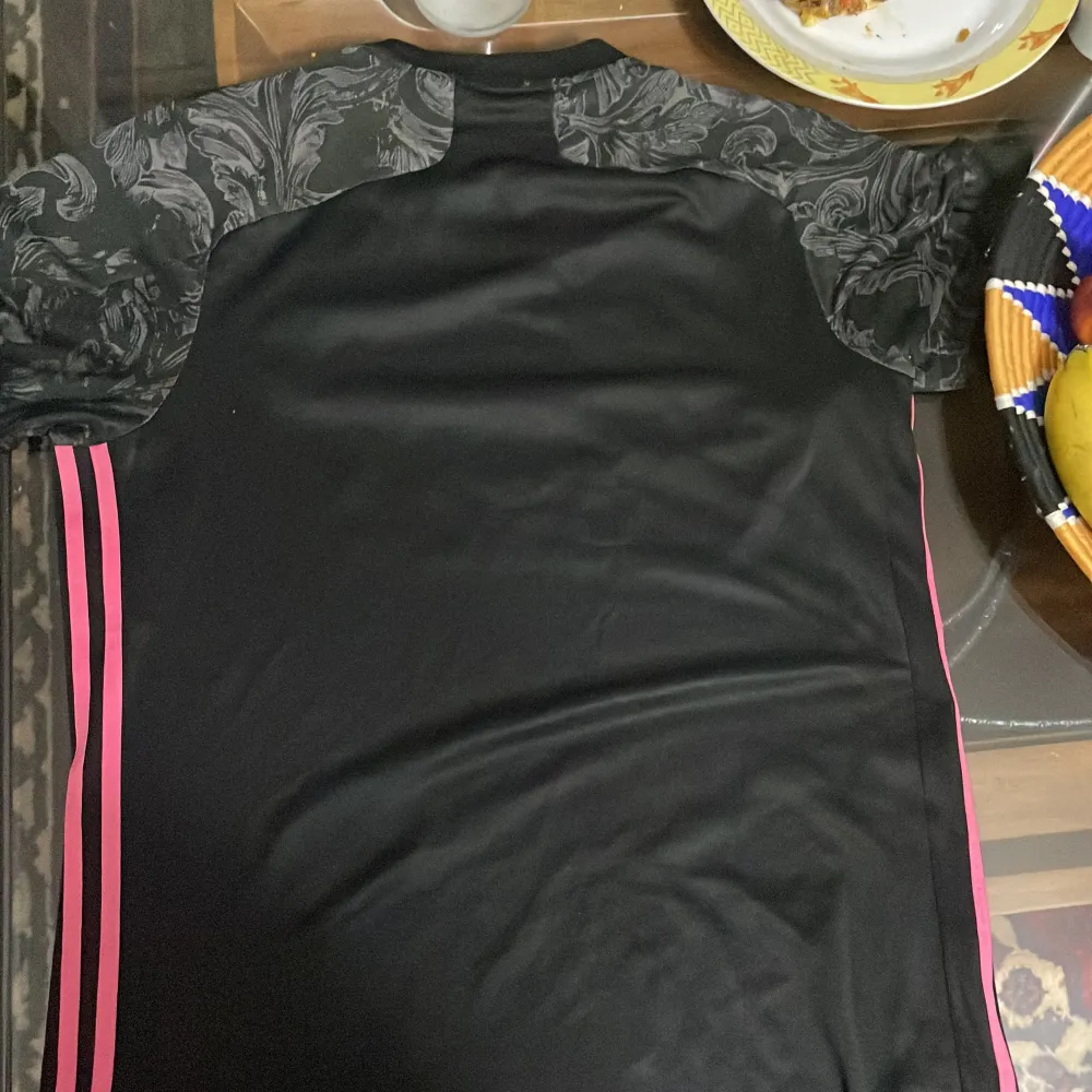 Säljer en snygg svart fotbollströja från Adidas med rosa detaljer och klubbmärke. Tröjan har korta ärmar och ett unikt mönster i tyget. Perfekt för fotbollsfans som vill ha något stilrent och bekvämt. AEROREADY-materialet håller dig torr och sval under matchen. Passa på att köpa denna coola tröja!. T-shirts.