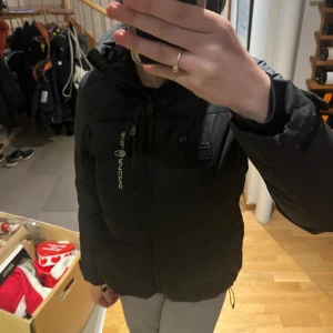 Svart dunjacka från Sail Racing - Säljer en svart dunjacka från Sail Racing i mycket bra skick. Jackan har en mysig huva och dragkedja framtill. Perfekt för kalla vinterdagar. Den är varm och bekväm, idealisk för både vardag och utomhusaktiviteter. Passar perfekt för vinterns alla äventyr! Inga defekter. Nypris 2500