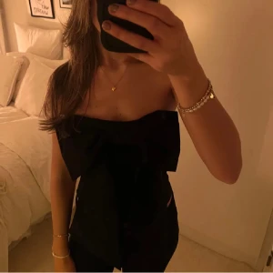 Svart axelbandslös topp med rosett - Snygg svart axelbandslös topp med en stor rosett framtill. Perfekt för en utekväll eller fest. Toppen har en elegant och stilren design som passar till många olika outfits.