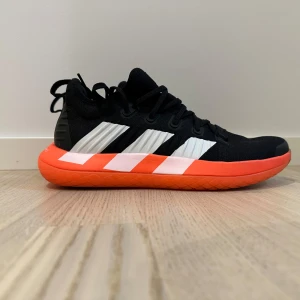 Svarta och orangea sneakers från Adidas perfekt för att spela tenis eller Padel  - Säljer ett par snygga Adidas sneakers i svart och orange. De har en cool design med vita detaljer och en skön, dämpad sula. Perfekta för träning eller vardagsbruk. Skorna är i nytt skick och har aldrig använts. Storlek och passform är normal.