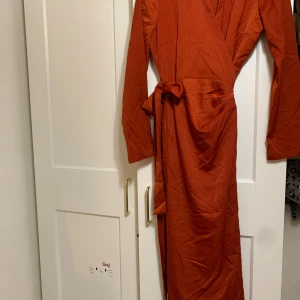 Rostfärgad omlottklänning - Säljer en snygg rostfärgad omlottklänning med långa ärmar. Klänningen har en knytning i midjan för en smickrande passform. Perfekt för höst och vår, och kan stylas både upp och ner beroende på tillfälle. Materialet känns mjukt och bekvämt mot huden.