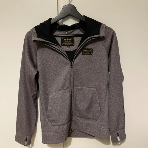 Grå hoodie från Burton - Säljer en snygg grå hoodie från Burton. Den har en dragkedja framtill och en stor huva. På ärmen finns det ett coolt tryck med 'BURTON' i svart. Jackan har långa ärmar med tumhål, vilket är perfekt för extra komfort. Den är i bra skick och passar perfekt för höst och vår.