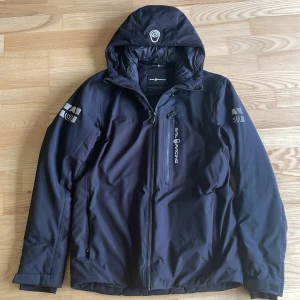 Svart jacka från Sail Racing - Säljer en svart jacka från Sail Racing. Den har en dragkedja framtill och en justerbar huva. Jackan är perfekt för kyligare väder och har snygga logodetaljer på ärmarna och huvan. Nypris är 3 000 kr men den är använd, däremot finns inga defekter