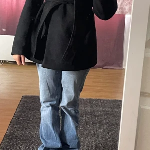 Svart kappa från Amisu - Säljer en stilren svart kappa från Amisu i storlek 36 (EU). Den har ett klassiskt snitt med knytband i midjan för en snygg passform. Perfekt för höst och vår, och funkar lika bra till vardags som till fest. Kappan är i bra skick och redo för en ny ägare!