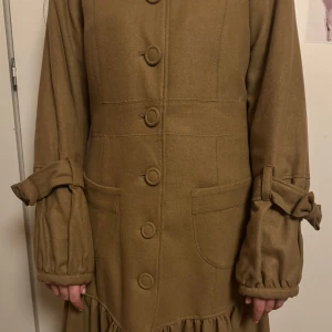 Beige kappa från S'NOB - Säljer en superfin beige kappa från S'NOB. Den har en klassisk stil med stora knappar framtill och söta knytband vid ärmsluten. Materialet känns mjukt och bekvämt, perfekt för höst & vinter. Kappan har en fin volangdetalj längst ner. För stor för mig