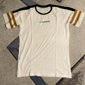 Vit t-shirt från Grunt - Säljer en snygg vit t-shirt från Grunt med texten 'G.HOMME' tryckt på bröstet. Den har korta ärmar med coola guldfärgade ränder och svarta detaljer vid kragen och axlarna. Perfekt för en avslappnad stil! Passar M/L