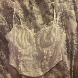 Vit spetskorsett med volanger - Säljer en superfin vit korsett i spets med volangdetaljer. Den har smala axelband och en söt, romantisk stil. Perfekt för fest eller en speciell kväll. Passar bra till både kjol och byxor!