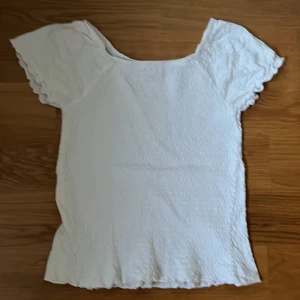 Vit topp från Lindex - Säljer en söt vit topp från Lindex. Den har korta ärmar med volangdetaljer och ett diskret blommönster. Materialet är stretchigt och ger en bekväm passform. Perfekt för sommardagar!