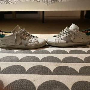 Vita sneakers med gröna detaljer - Säljer ett par vita golden goose med gröna detaljer. De har en klassisk design med snörning och en stjärna på sidan. Skorna är i skinn och har en lite sliten look som ger dem en cool vintage känsla. Perfekta för både vardag och fest!