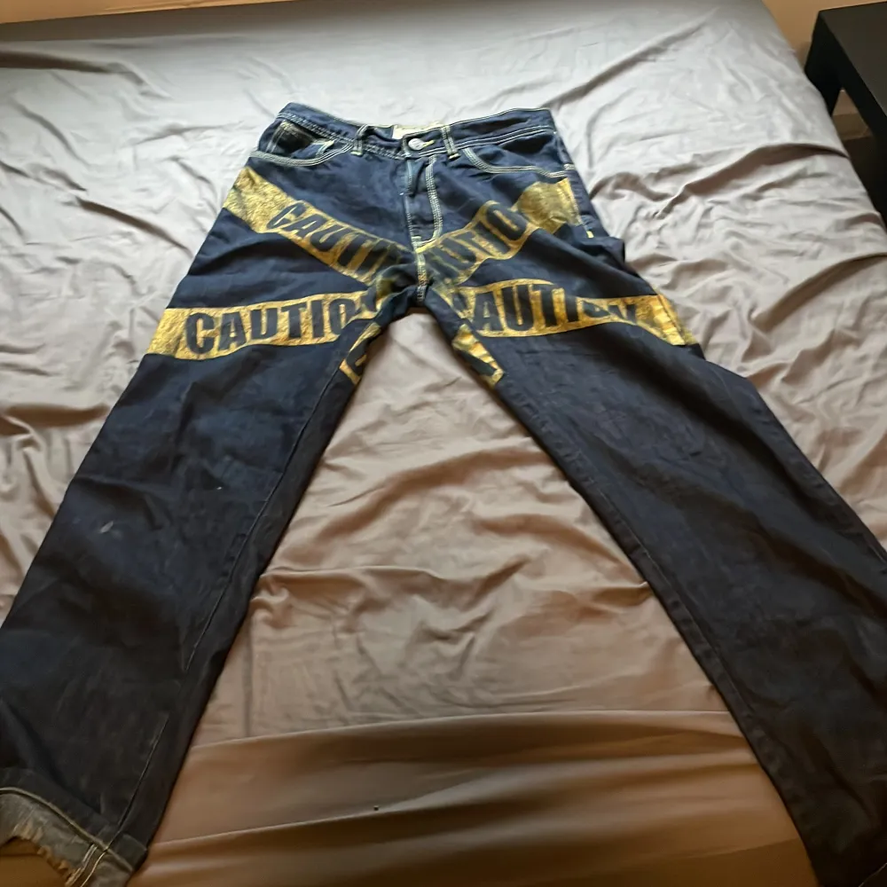 Säljer ett par unika jeans med coolt CAUTION tryck i gult över hela byxorna. De är mörkblå och har en baggy passform. Perfekta för dig som vill sticka ut och göra ett statement med din stil! Liten fläck synat inte mkt försvinner förmodligen i tvätten. Jeans & Byxor.